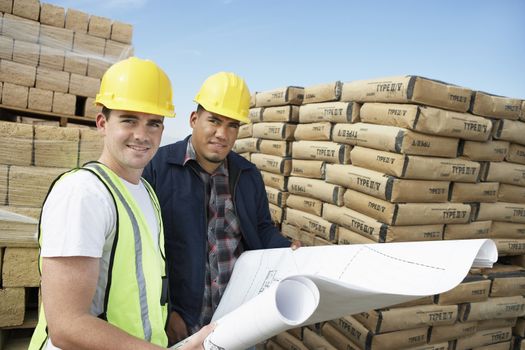 Portrait of two workers with plans in front of cement bags and bricks