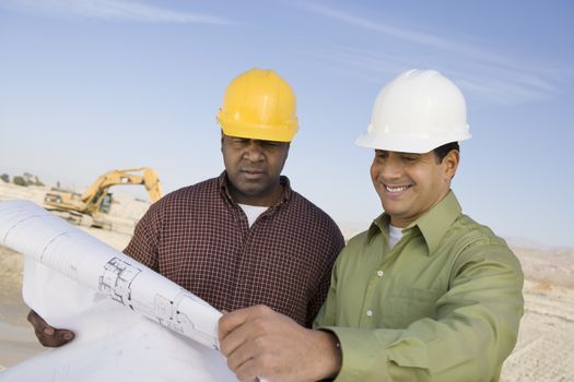 Two multiethnic construction workers with blueprints against the sky at site