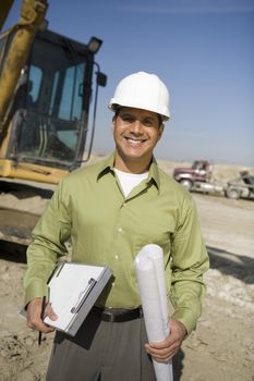 Portrait of a happy foreman standing with clipboard and blueprint at construction site