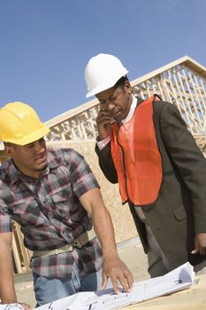 Architect using phone with co-worker standing with blueprint at construction site