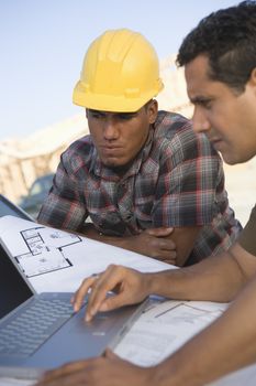 Mature man working on laptop with co-worker besides at construction site