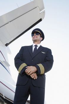 Low angle view of a smiling pilot in uniform standing beside an airplane with hands clasped 