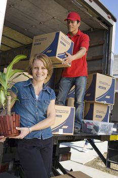 Portrait of happy woman holding pot plant with delivery man unloading moving boxes from truck