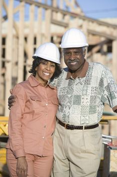Portrait of happy senior African American couple wearing hardhat in front of incomplete house