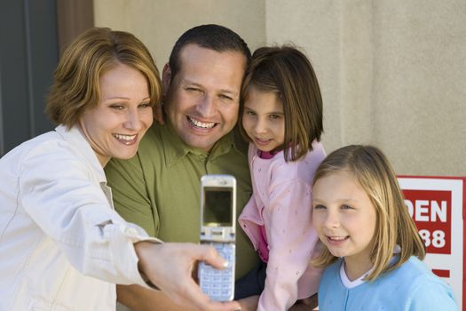 Happy family of four taking self-portrait through cell phone in front of new house