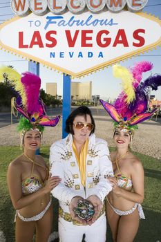 Portrait of Elvis Presley impersonator holding chips and standing with casino dancers against sign board