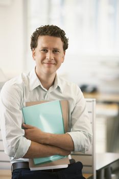 Portrait of happy young businessman with files standing by cubicle
