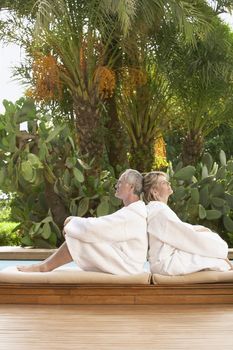 Side view of middle aged couple in bathrobes sitting back to back by pool