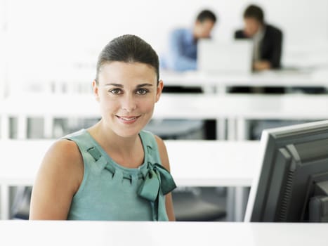 Portrait of happy young businesswoman at computer desk with male colleagues in background