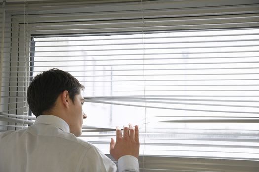 Rear view of a businessman looking out of the office window through blinds