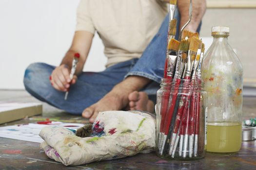 Low section of a male artist sitting on floor with paint brushes and materials