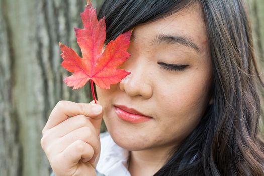 Autumn portrait of cute young woman in front of a maple tree holding a maple leaf and covering her eye