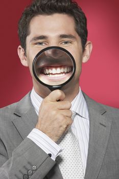 Closeup of a businessman holding magnifying glass to smiling mouth against red background