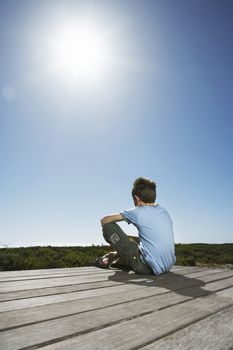 Rear view of young boy sitting on boardwalk against clear sky