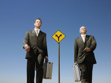 Confident businessmen with briefcases walking past road sign against blue sky
