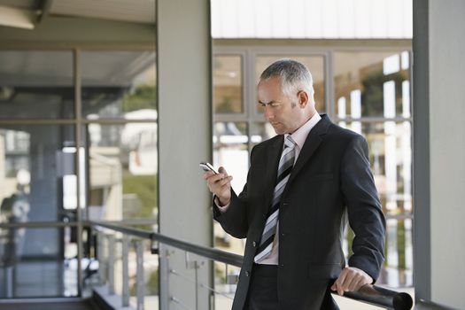 Middle aged businessman leaning on railing while using mobile phone in office