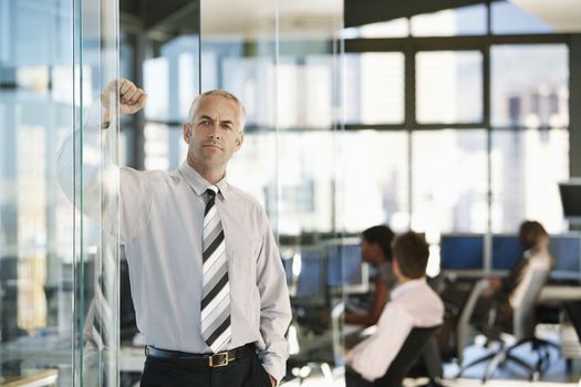 Confident middle aged businessman leaning on glass door with colleagues working in background