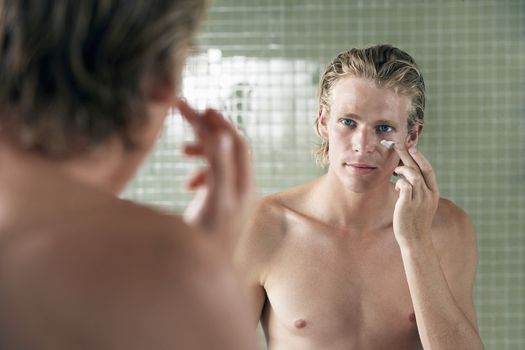 Handsome young man applying facial cream in front of mirror