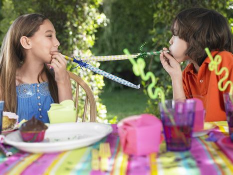 Young boy and girl blowing party puffers at each other in outdoor birthday party