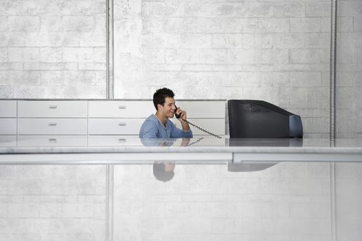 Young businessman sitting at computer desk using landline phone in office