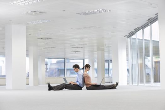 Side view of businessman and businesswoman sitting back to back using laptops in empty office space