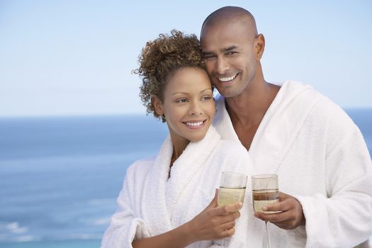 Couple in bathrobes holding drinks embracing ocean in background half length