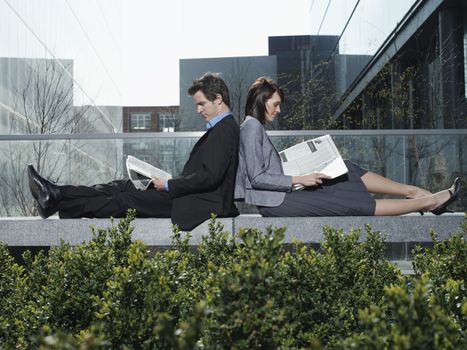 Side view of young businessman and businesswoman reading newspapers while sitting back to back on wall