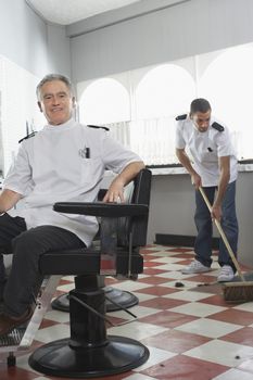 Portrait of happy senior male barber with apprentice sweeping floor at barbershop