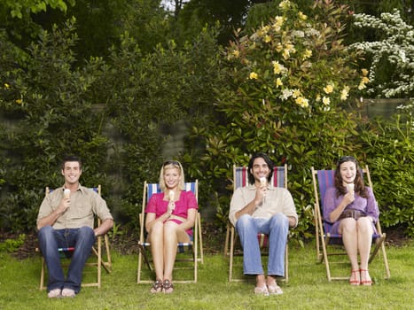 Full length group portrait of young people sitting in row with icecreams on deckchairs in garden