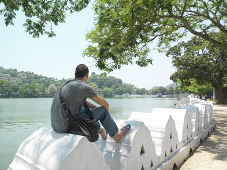 Young man sitting on surrounding wall by river back view