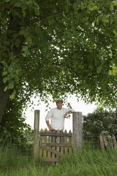 Portrait of a smiling man standing across field gate under the tree