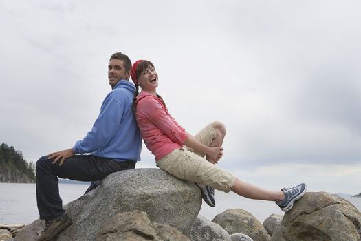 Full length side view of a happy young couple sitting back to back on rocks against ocean