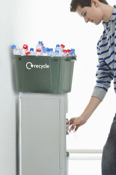 Young man filling glass of water from cooler with recycle bin on it