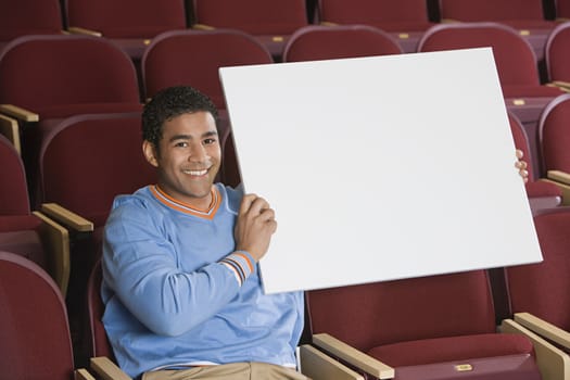 Portrait of happy African American man in casual wear holding a blank placard while sitting in auditorium