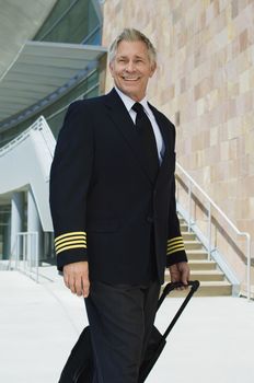 Portrait of a smiling senior airline pilot pulling suitcase outside building