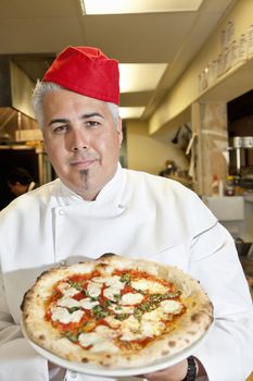 Portrait of a confident chef wearing headgear while holding pizza in commercial kitchen