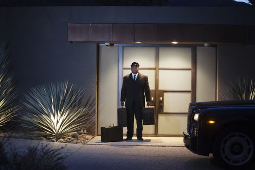 Chauffeur stands at lit entrance doorway with luggage