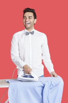 Cheerful man in formals ironing shirt over red background