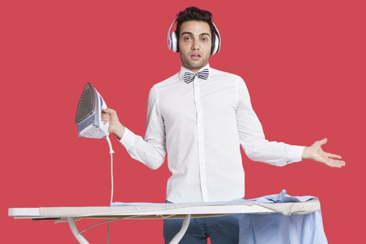 Portrait of a confused man in formals ironing as he listens to music over red background