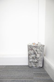Wastebasket full of crumpled paper in corner against white wall