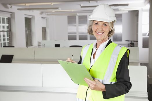 Portrait of smiling senior woman in reflector vest and hard hat holding clipboard at office