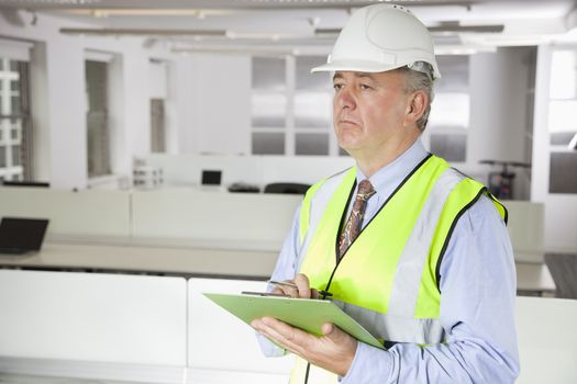 Middle-aged man in reflector vest and hard hat with clipboard at office
