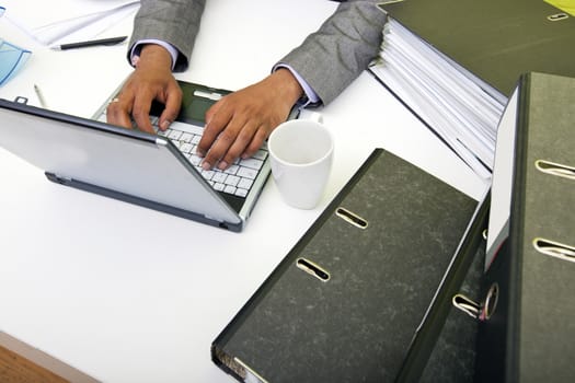 Close up of Indian mans hands typing on laptop with folders and mug