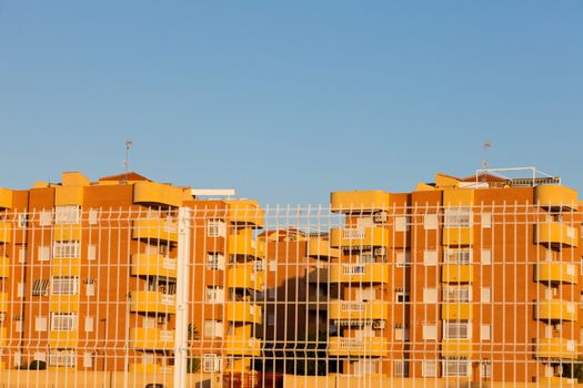 Group of multistorey colorful yellow-orange modern architecture apartment blocks behind security fence in sunshine of Spain, Europe, against clear blue sky