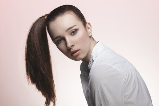 pretty young woman with brown creative hair-style posing with ponytail on the right, wearing white shirt and looking in camera