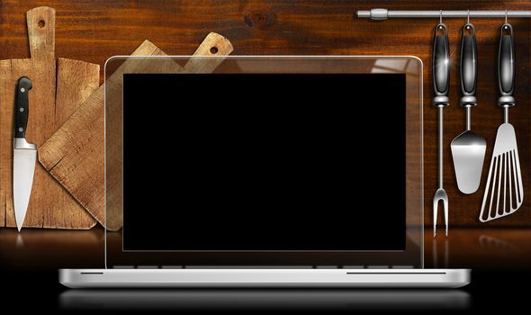 Laptop computer with black and transparent screen in a kitchen with cutting boards and utensils on wooden wall. Template for recipes or food menu