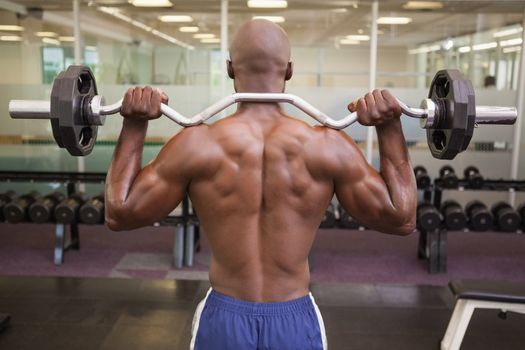 Rear view of a shirtless muscular man lifting barbell in gym