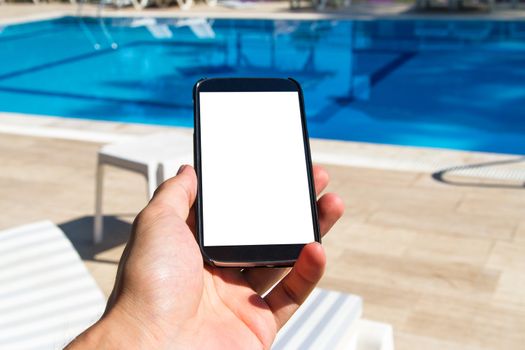 Hand holding blank, white, empty screen smart phone with copy space on poolside.