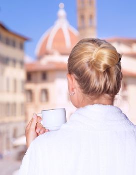 Back side on woman having morning coffee in the hotel, wearing white robe and enjoying cityscape from the window, Europe, Italy, Florence