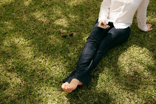 High angle view of young woman in jeans and white shirt resting in city park, sitting on grass and text messaging with mobile phone. Copy space, cropped view
